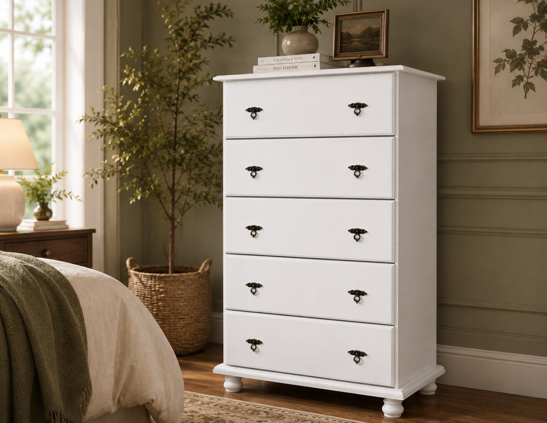 A large white five-drawer chest of drawers with dark metal knobs, styled in a bedroom next to a bed and potted plant.