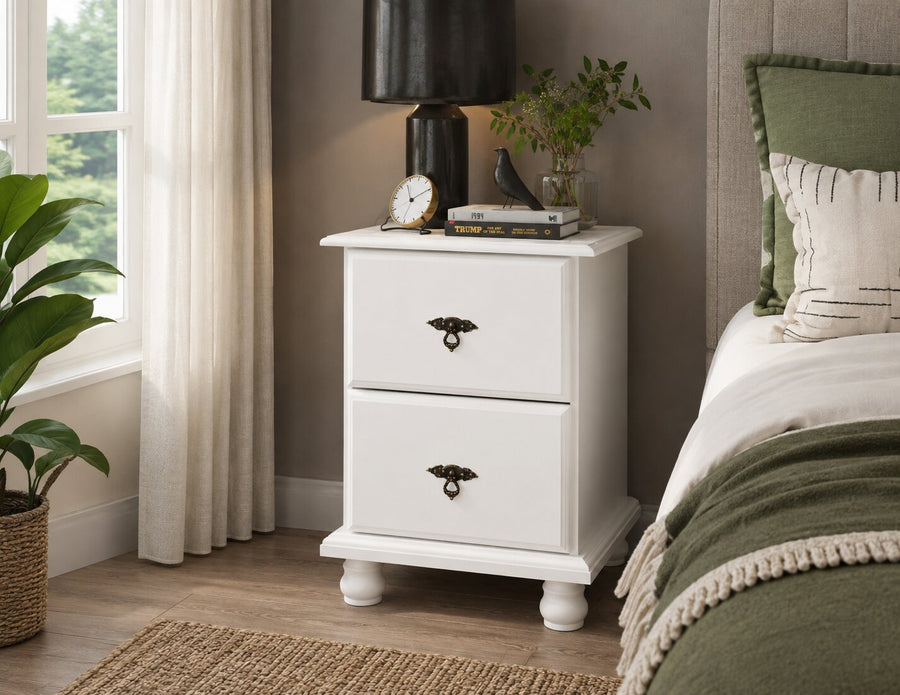 A white two-drawer nightstand with dark metal hardware, styled in a bedroom next to a bed with green bedding and a window with sheer curtains.