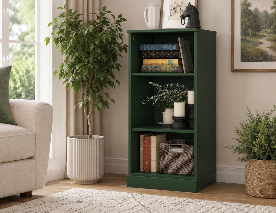 A tall, dark green three-tier bookcase styled in a cozy living room corner next to a window, potted plants, and a beige armchair.