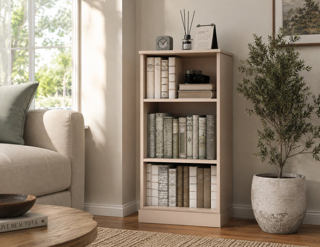 A tall, narrow three-tier bookcase in a light beige finish stands in a styled living room corner next to a sofa and a potted plant. The bookcase is styled with books, a small clock, a diffuser, and a picture frame.