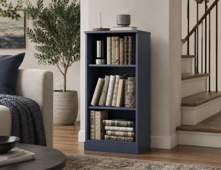 A tall, narrow antique blue bookcase with three open shelves, styled with vintage books and small decor items, set in a cozy living room next to a staircase.