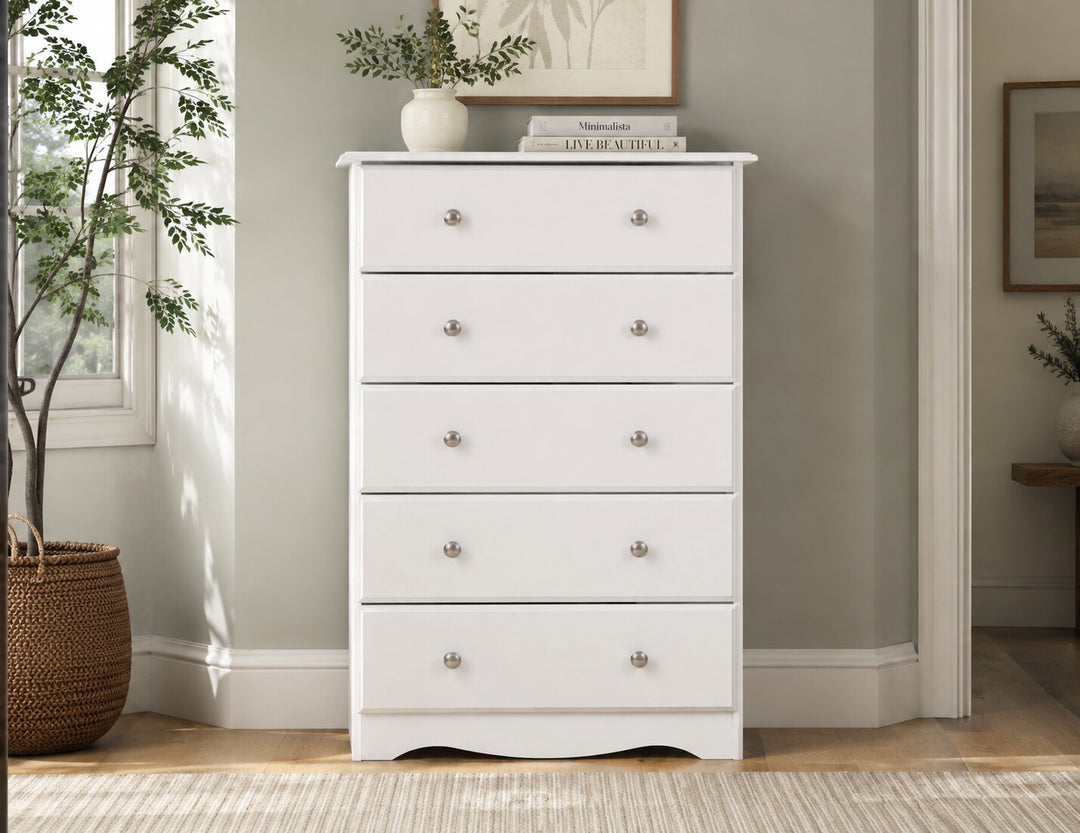 A white five-drawer chest of drawers with small round silver knobs, styled in a bright room with a potted plant, books, and framed wall art.