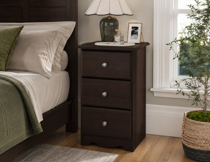 A three-drawer dark brown wooden nightstand with silver knobs, styled in a bedroom next to a bed with white linens and a green throw blanket. The nightstand holds a table lamp and a small picture frame, set on a light wood floor next to a potted plant.