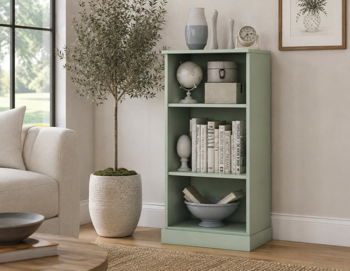 A sage green 3-shelf bookcase styled with books, vases, and a globe in a bright living room next to a beige sofa and a potted tree.
