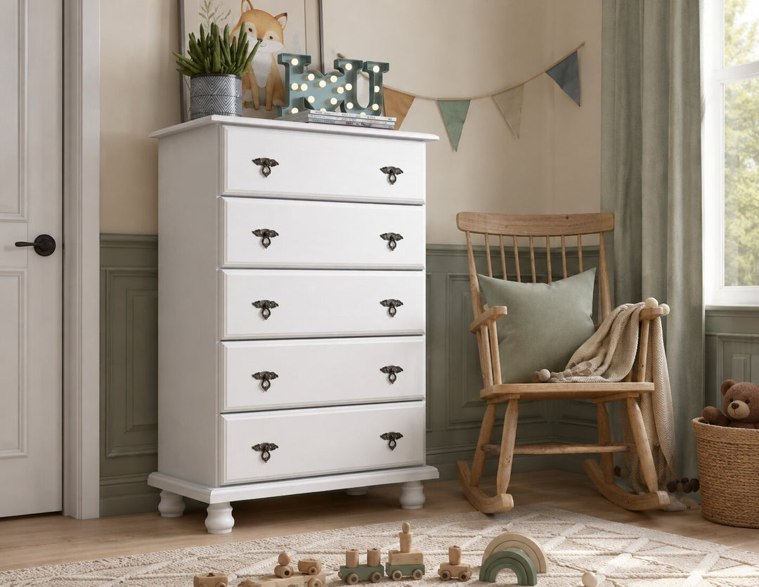 A tall white five-drawer dresser with dark metal handles stands in a styled nursery or playroom corner next to a wooden rocking chair, with toys on the floor and a window in the background.
