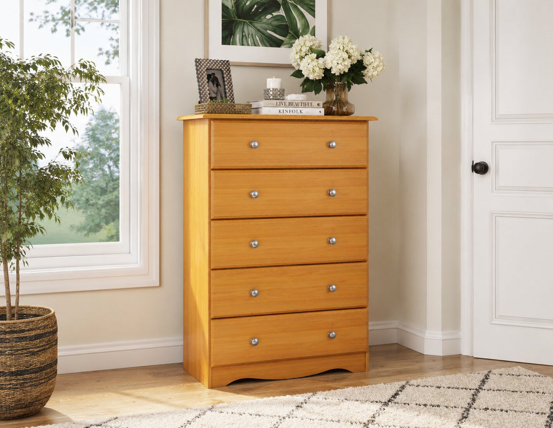 A tall five-drawer wooden chest of drawers in a warm honey pine finish, styled in a bright room next to a window and potted plant.
