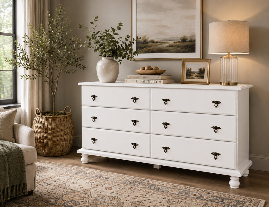 A white wooden dresser with 6 drawers and dark metal handles, styled in a cozy, warm-toned room next to a window, potted plant, and a matching table lamp.