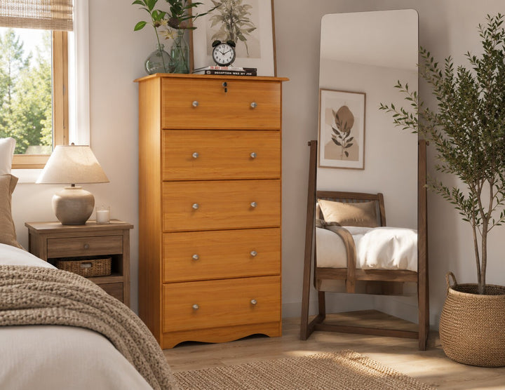 A tall wooden five-drawer dresser in a warm honey finish stands in a styled bedroom next to a bed, with a full-length mirror, bedside lamp, and potted plant in the background.