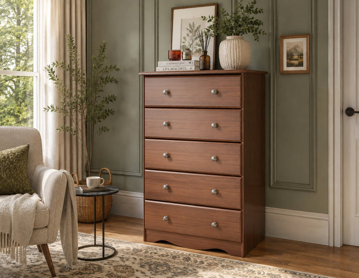 A five-drawer wooden chest of drawers in a warm medium brown finish, styled in a cozy room with sage green paneled walls, a beige armchair, and a patterned rug.